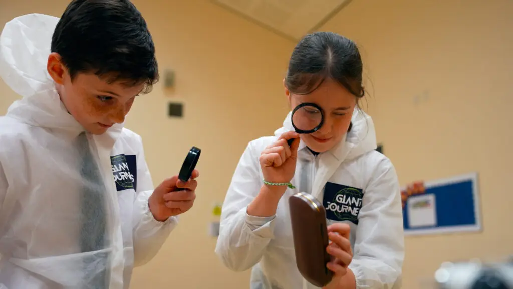 Two children in white lab coats investigate objects using magnifying glasses indoors.
