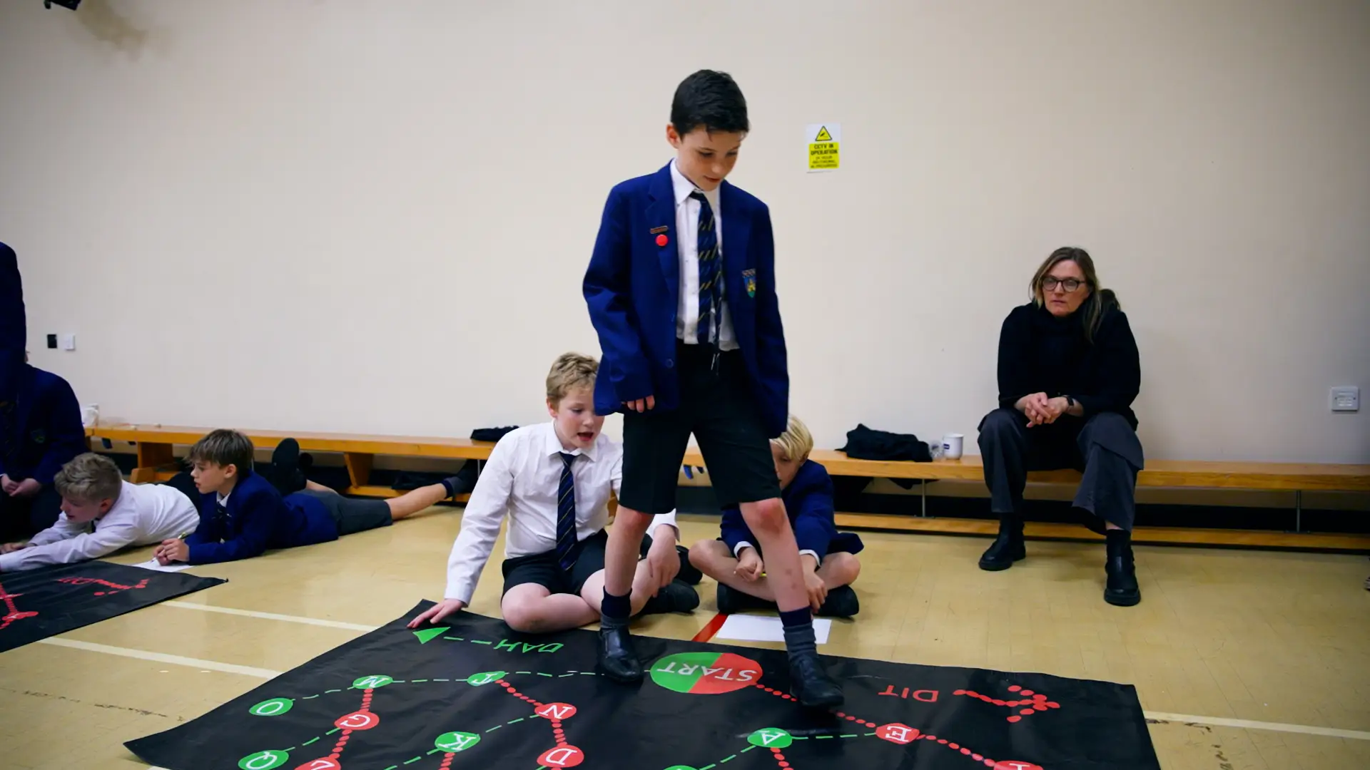 Boys in school uniforms engage with interactive floor mats in a classroom, while a woman observes.