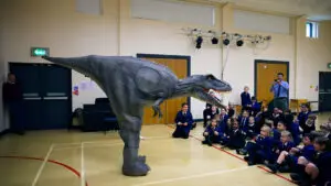 A large dinosaur model stands in a school hall as children sit excitedly in front.