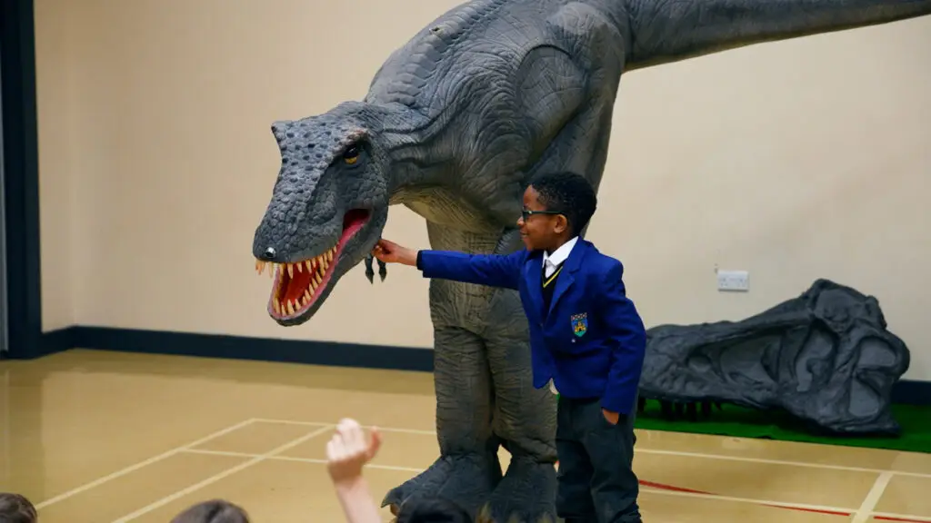 Child in a blue jacket interacts with a large dinosaur model in a school classroom setting.