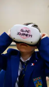 Child wearing a virtual reality headset labeled "GIANT JOURNEYS" in a classroom setting.