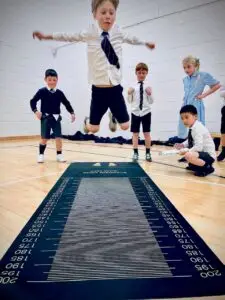 Boy jumping on a long jump mat while four peers observe in a gymnasium.