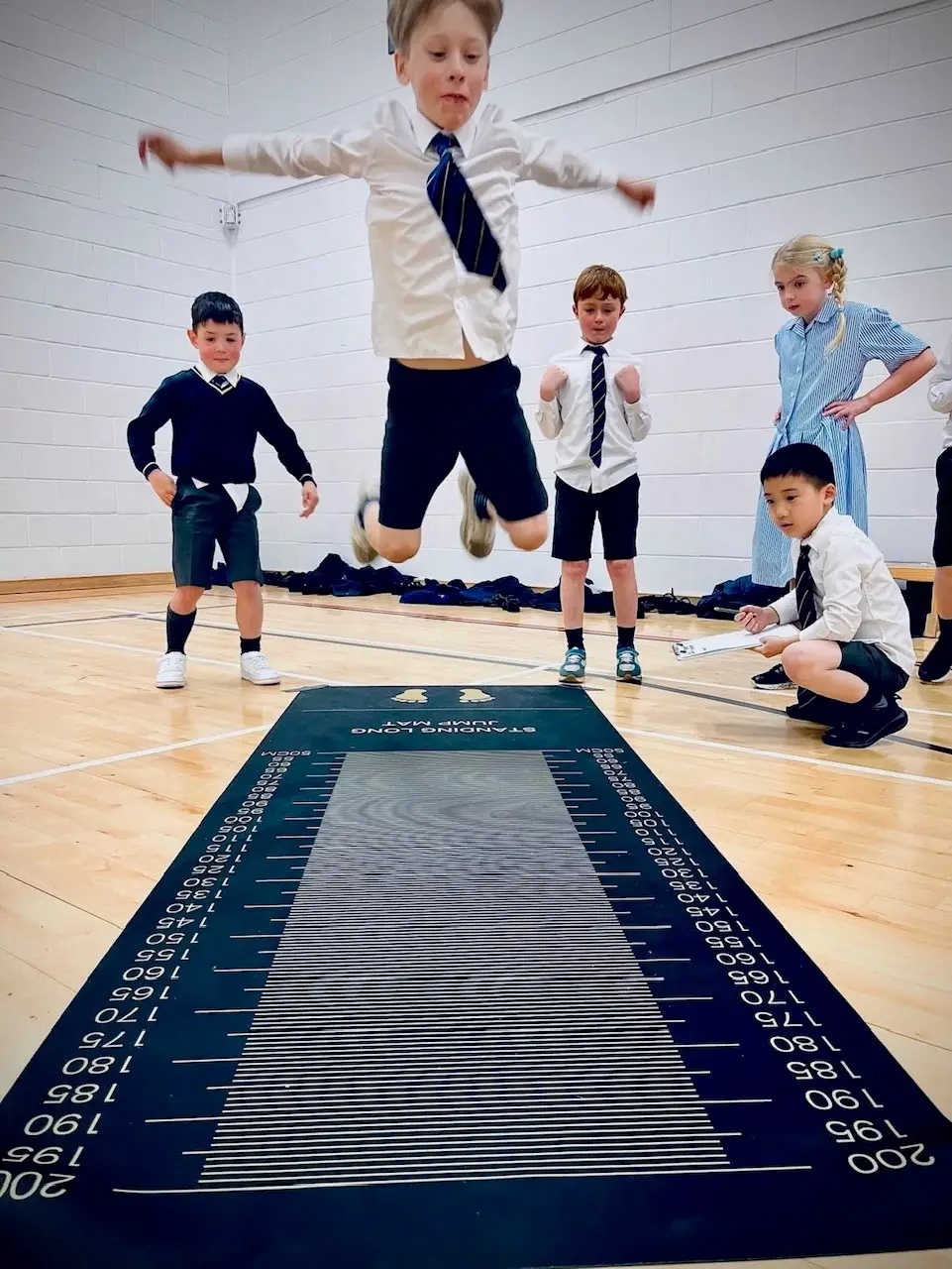 Boy jumping on a long jump mat while four peers observe in a gymnasium.