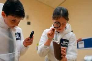 Landscape Still 01 Two children in white lab coats examining objects with magnifying glasses indoors.