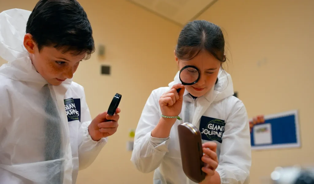 Two children in white lab coats investigate objects using magnifying glasses indoors.