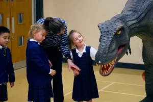 Landscape Still 04 Children interact with a large, lifelike dinosaur model in a classroom setting.