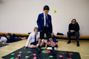 Landscape Still 07 Boys in school uniforms engage with interactive floor mats in a classroom, while a woman observes.