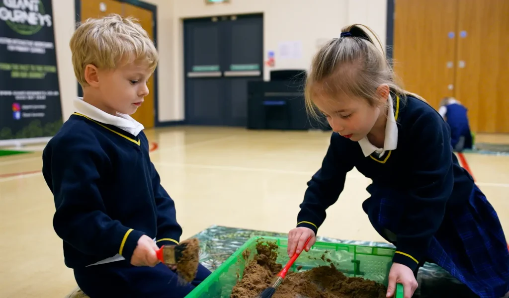 Two children in school uniforms dig in sand in a green container, using small red tools.