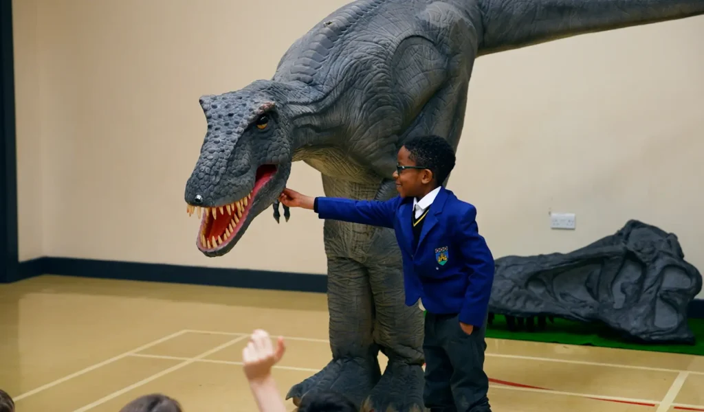 Child in a blue jacket interacts with a large dinosaur model in a school classroom setting.