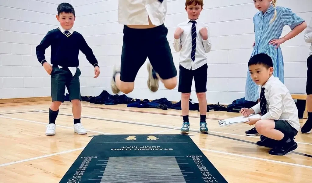 Children in school uniforms participate in a long jump activity on a mat.